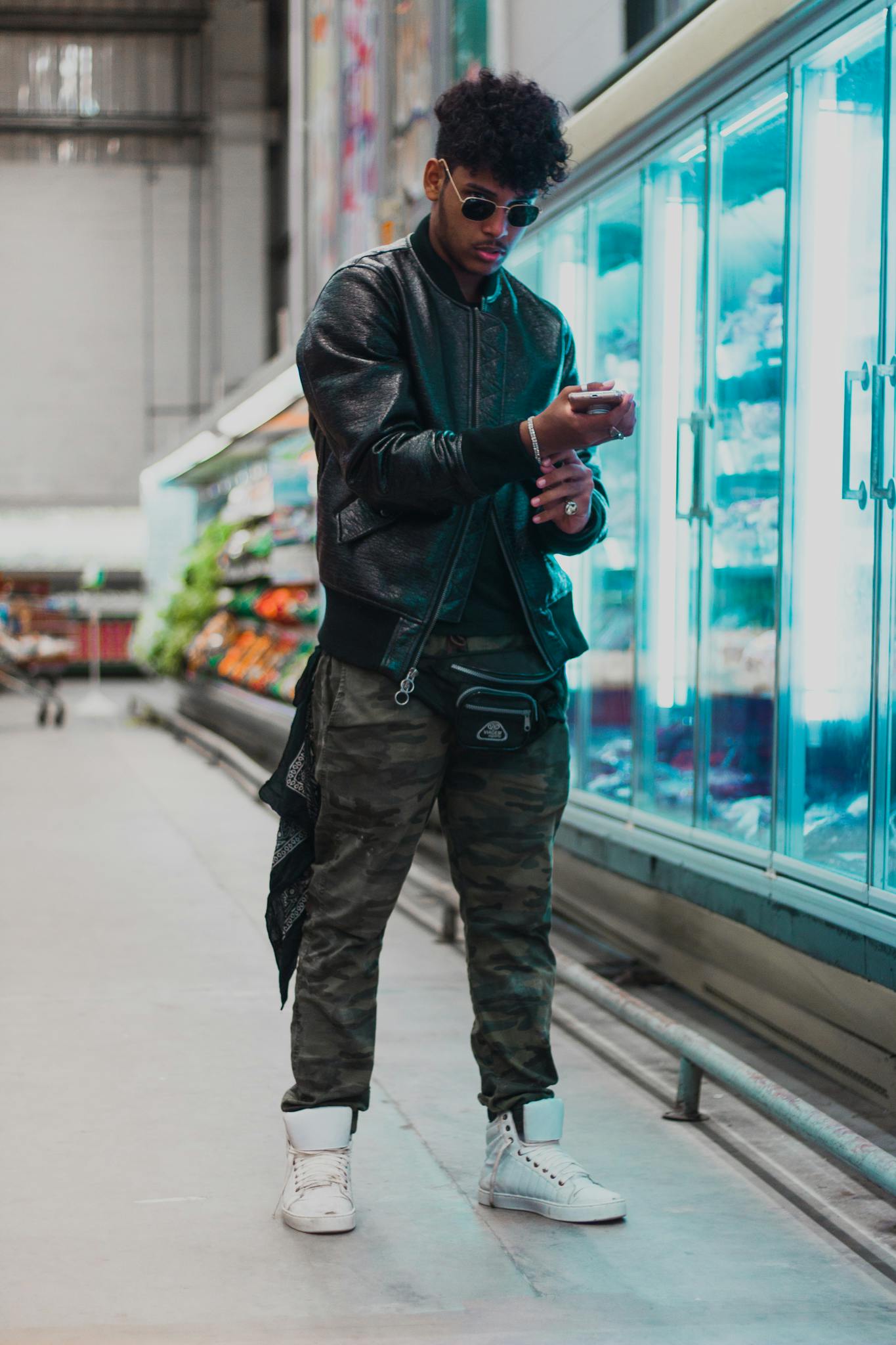 Fashionable young man in leather jacket and camo pants checking phone at grocery store.